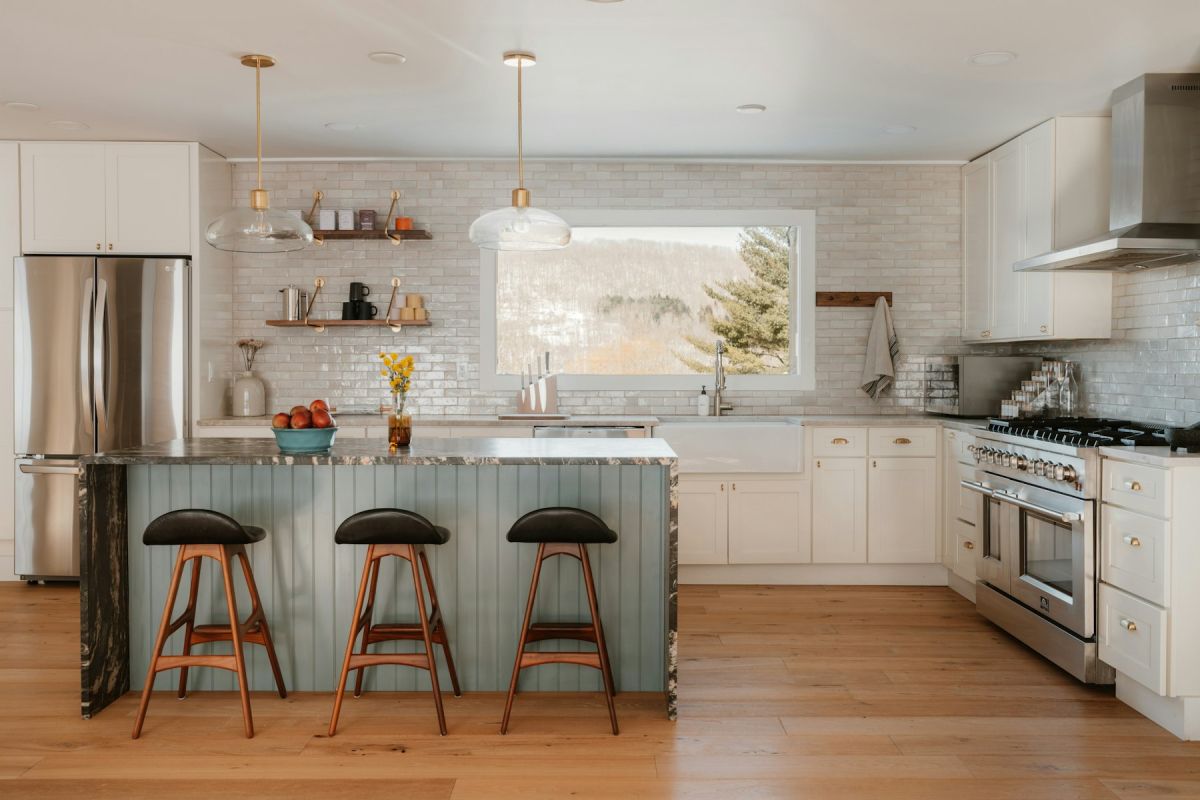Bright kitchen with stone-topped island, leather stools, white tile backsplash, and stainless range with hood